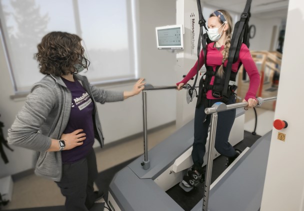 A physical therapist standing next to a patient who is strapped into a harness and stepping device.