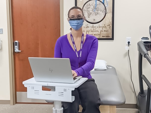 Danielle wearing a surgical mask next to a physical therapy table and a computer.