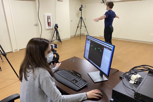 A male research participant wearing sensors stands in the middle of a room with arms stretched out wide while a female researcher monitors data on a computer screen.