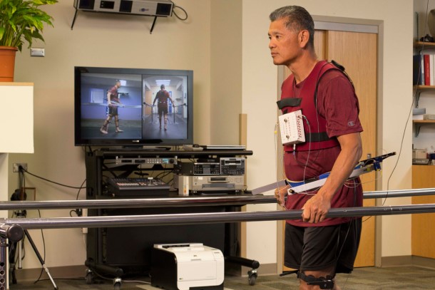 A male patient wearing sensors walks down a runway while holding onto guard rails.