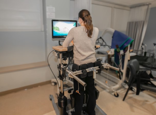 A woman stands leaning against a table with a computer monitor with help of a standing frame.