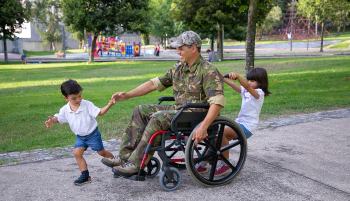 Soldier dressed in military uniform sits in wheelchair while children play with him.