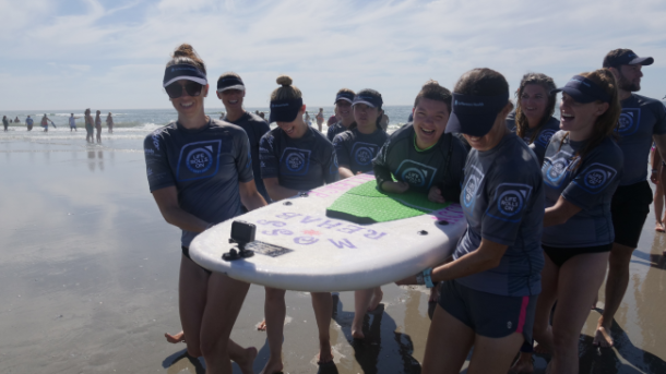 A group of volunteers carries a surfboard with a child riding on top.