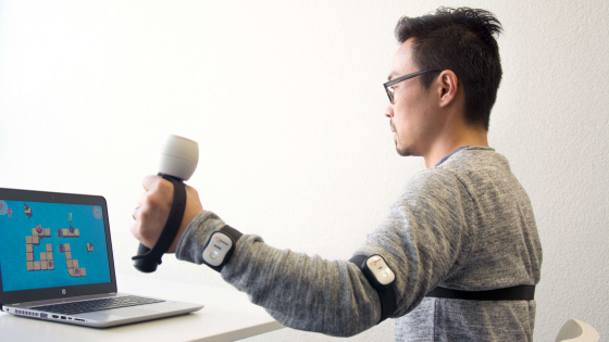 A male sitting in front of a laptop holding a baton in his left hand and sensors on his bicep and wrist.