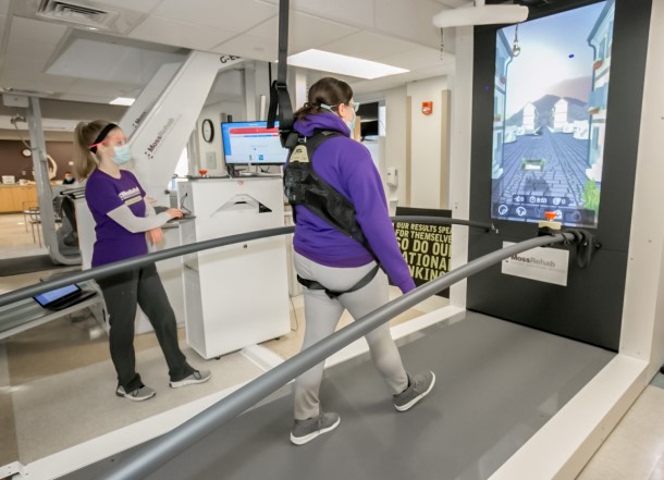 A woman being held up by a harness walks on a large treadmill while watching a LCD screen